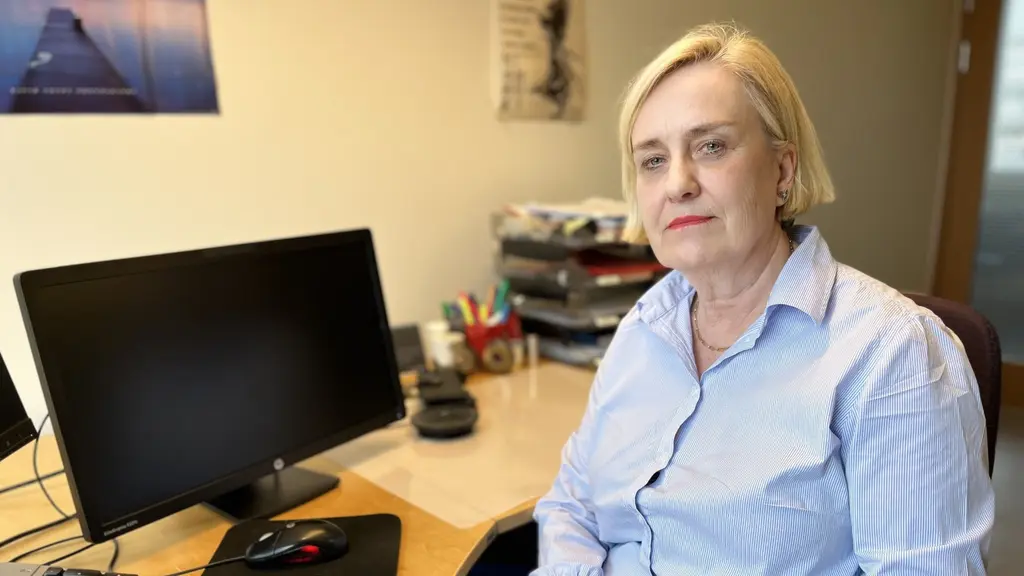 En person i randig skjorta sitter vid ett skrivbord med en datorskärm, tangentbord och kontorsmaterial. Bakgrunden visar en ljus vägg med tavlor och en dörröppning.