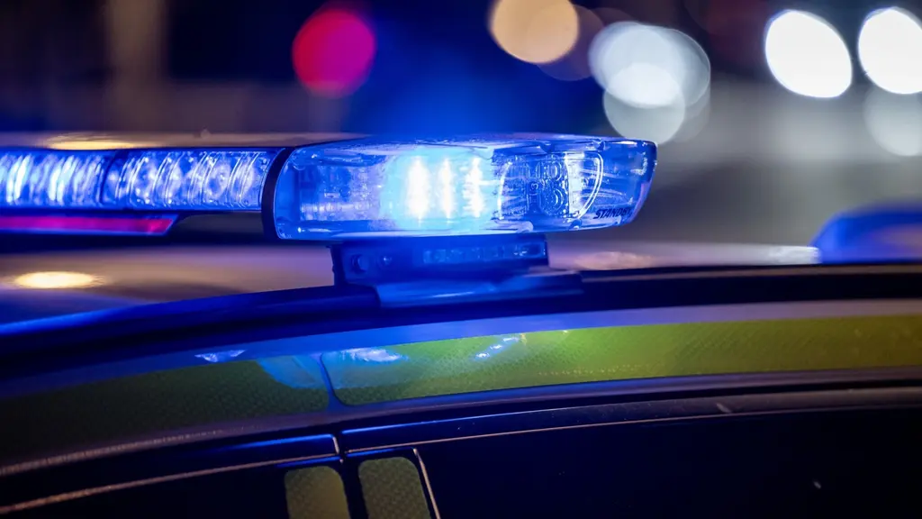Blåljus från en polisbil lyser i mörkret. Bilden visar en närbild av taket på en polisbil med blåljusen aktiverade, vilket skapar en dramatisk stämning. Bakgrunden är suddig med ljuspunkter.