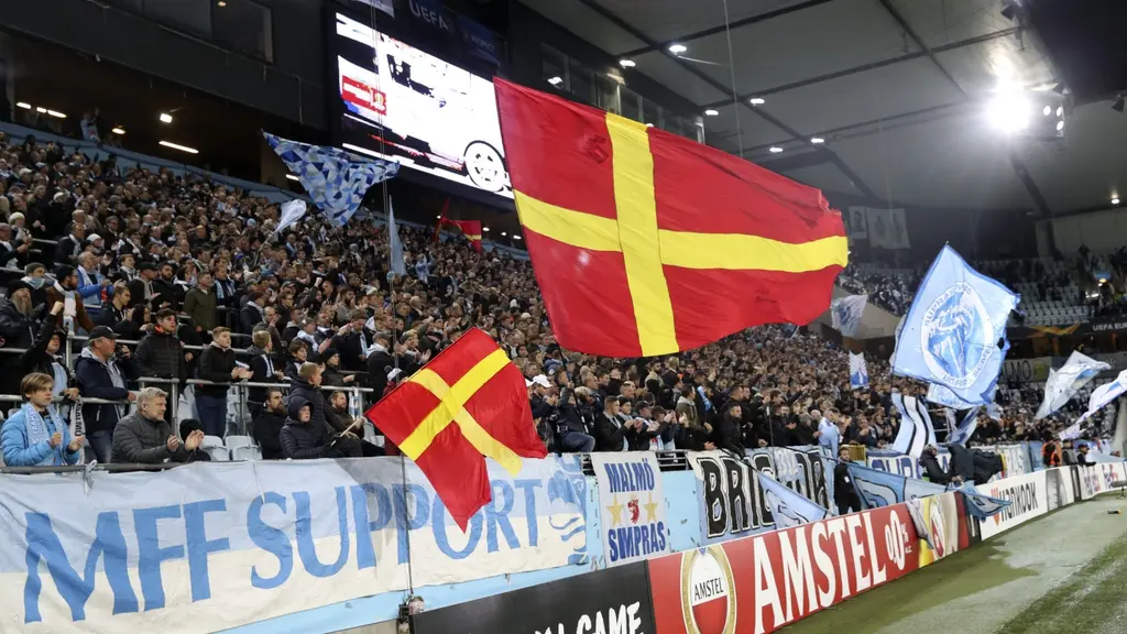 MFF-supportrar med flaggor och banderoller på läktaren inför Europa League-matchen mellan Malmö FF och FC Köpenhamn på Malmö stadion. Stämningen är livlig med färgglada flaggor och högljudda hejarop.