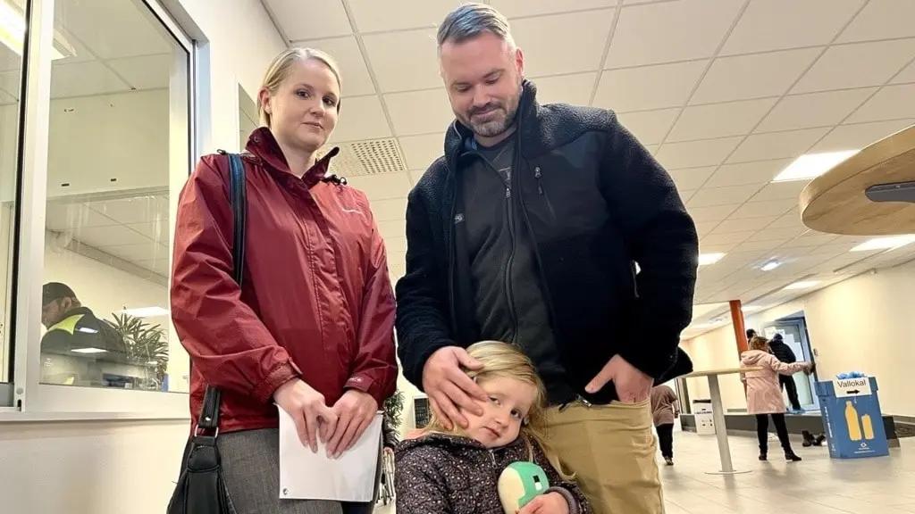 En vuxen person i röd jacka och en annan i svart jacka står bredvid ett barn som håller en mjukisdjur, inomhus i en offentlig miljö med människor och möbler i bakgrunden.