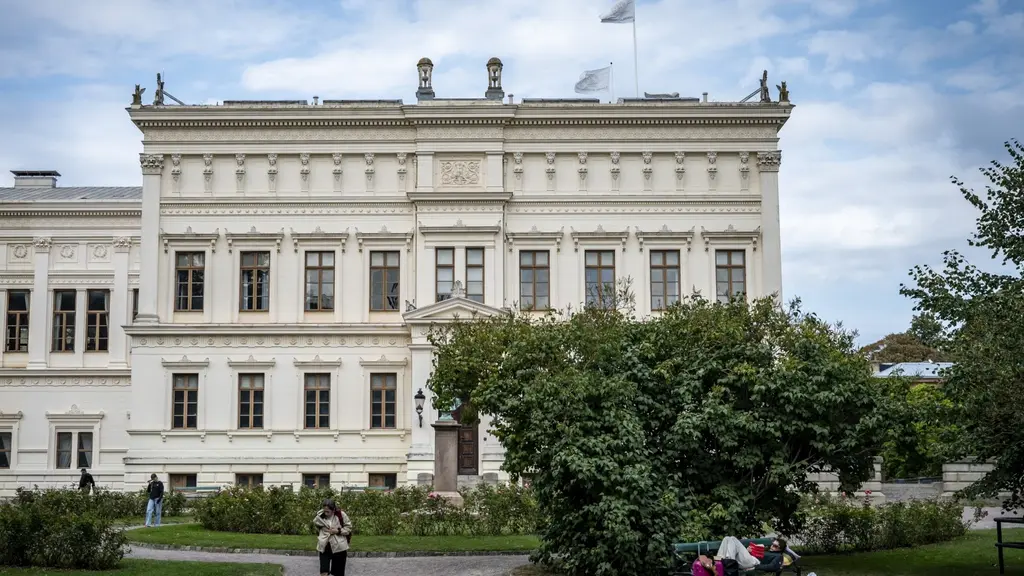 Universitetshuset vid Lunds universitet syns i bakgrunden med klassisk arkitektur. Framför byggnaden sitter människor på parkbänkar i Lundagård, omgivna av grönska och gångstigar.