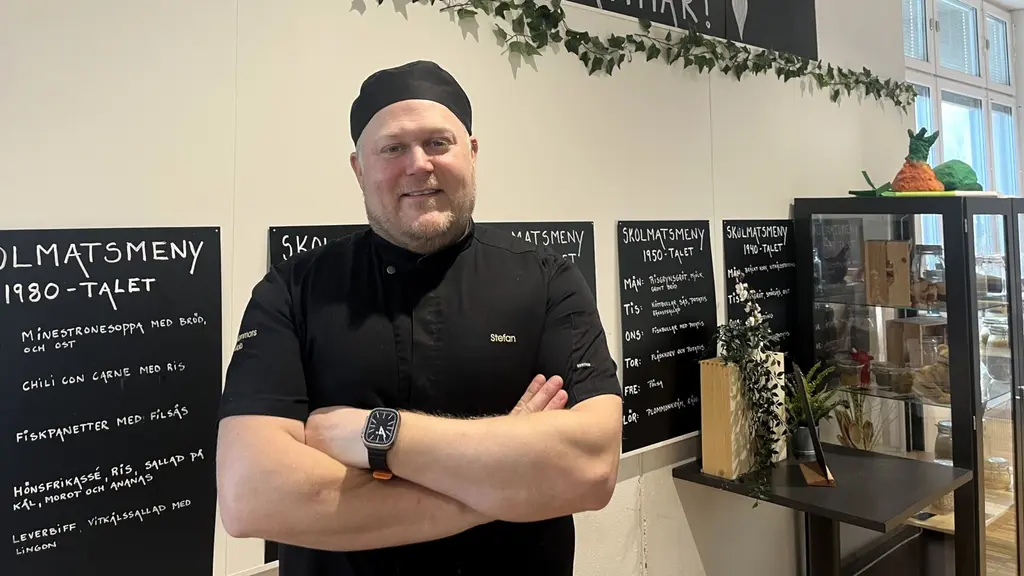 Skolkocken står i matsalen. Han är iklädd svart skjorta och mössa. Han har armarna i kors och ler mot kameran.