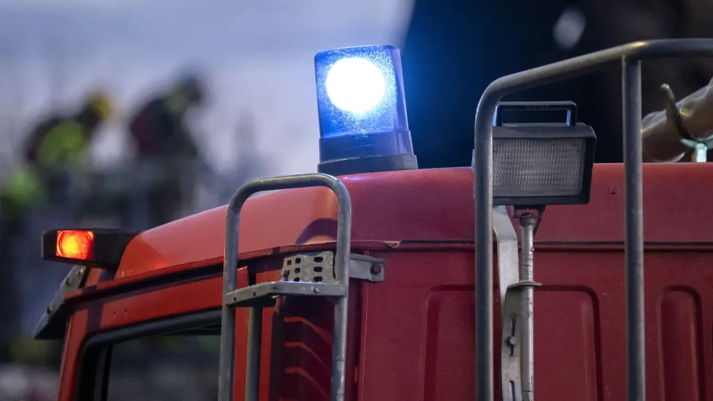 En röd brandbil med blåljus syns i förgrunden, medan räddningspersonal arbetar i bakgrunden. Det är kväll och himlen är mörk, vilket förstärker känslan av en pågående insats.