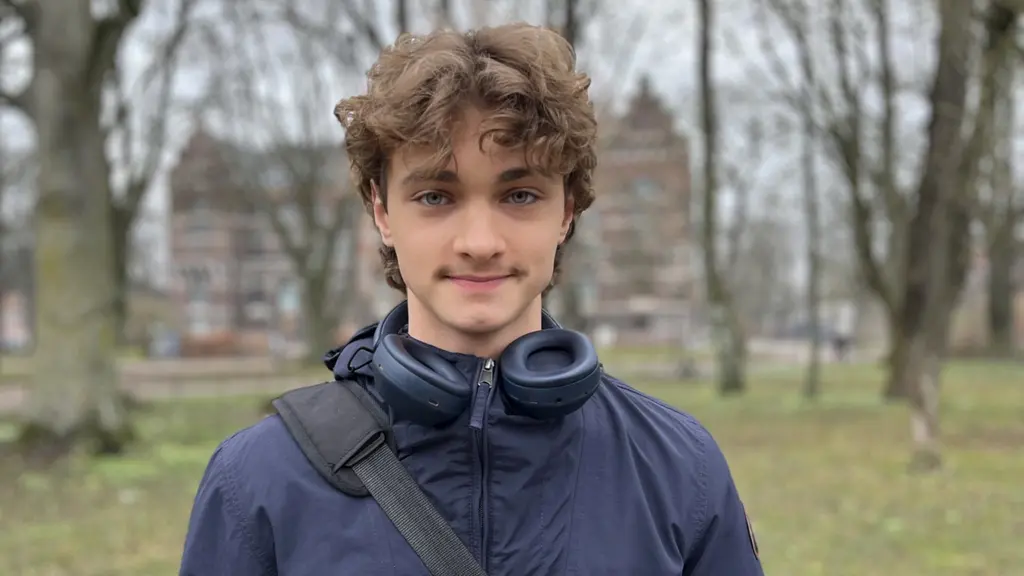 Ung kille med brunt lockigt hår och blå jacka står framför några träd och ser in i kameran. 