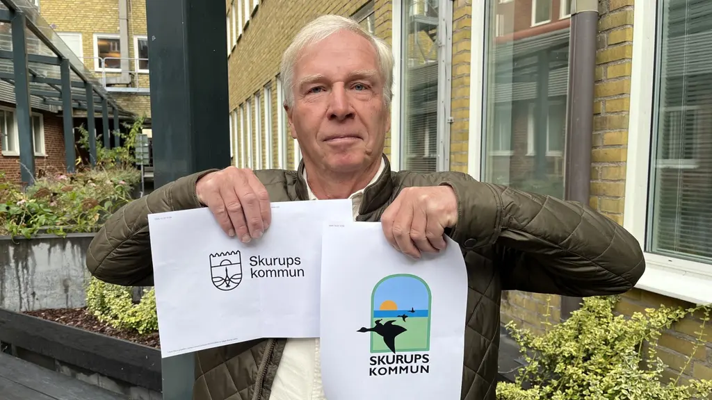En person håller upp två papper med Skurups kommuns gamla logotyp till höger och den nya logotypen till vänster. Bakgrunden visar en tegelbyggnad och grönska utomhus.