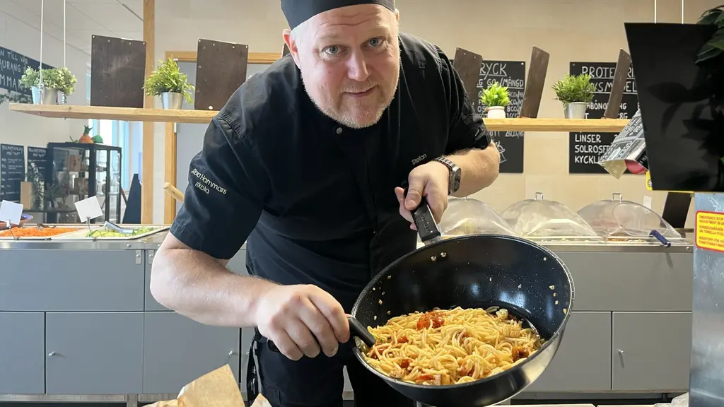 En kock klädd i svart uniform tillagar pasta i ett kök med en modern inredning. Kocken håller en stekpanna med pasta och grönsaker, omgiven av hyllor med gröna växter.