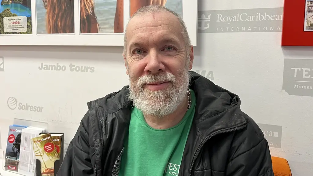 En person sitter inomhus framför en vägg med affischer och reklam. Personen bär en grön t-shirt och en mörk jacka. På bordet finns broschyrer och en kaffekopp.