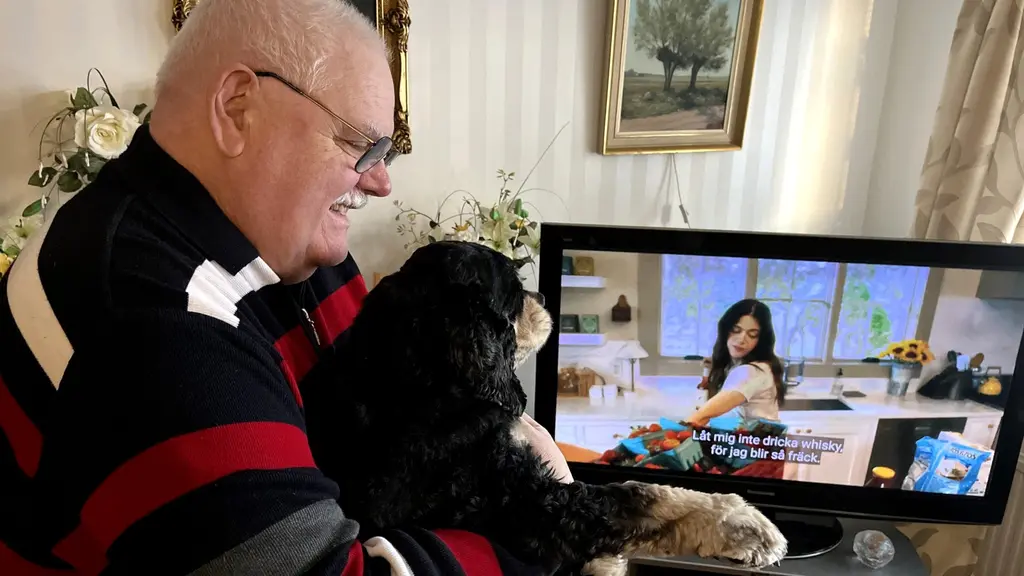 Husse Karl-Lennart Säll håller i sin hund framför en tv. Karl-Lennart ler mot hunden Pumba som har fullt fokus på tv:n.