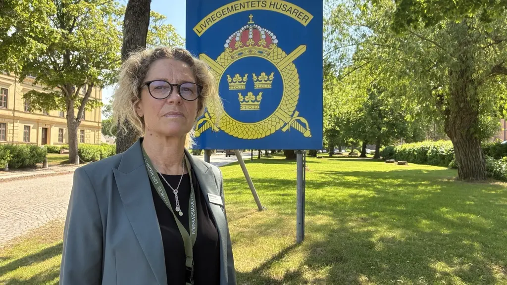 Birgitta Holm från K3 står utomhus framför en skylt i en grön parkmiljö.