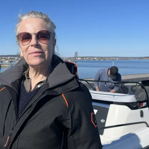 En person står vid en småbåtshamn i Mariestad med en motorbåt i bakgrunden. Soligt väder och klarblå himmel. Vattnet är stilla och omgivningen visar flera båtar förtöjda vid bryggor.