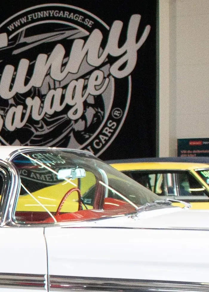 Två amerikanska veteranbilar står i Funny Garage-lokalerna utanför Skövde, framför en stor svart väggskylt med företagsnamnet.