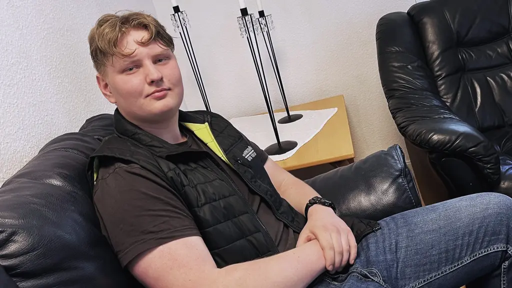 Hugo Johansson sitter i en svart skinnfåtölj i ett vardagsrum, klädd i svart väst och jeans.