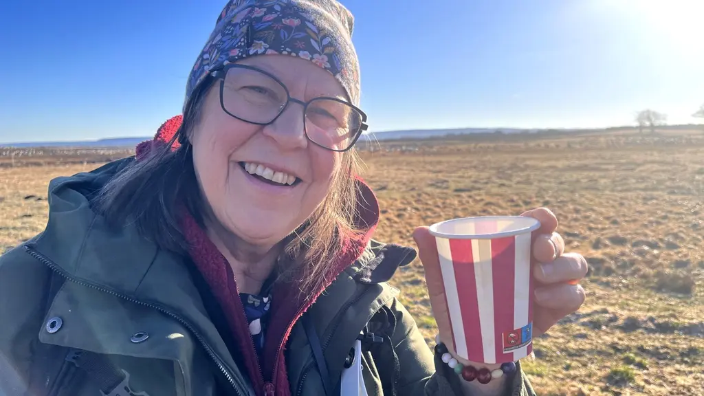 En kvinna med mössa, glasögon och en kopp kaffe vid en betesmark med fåglar.