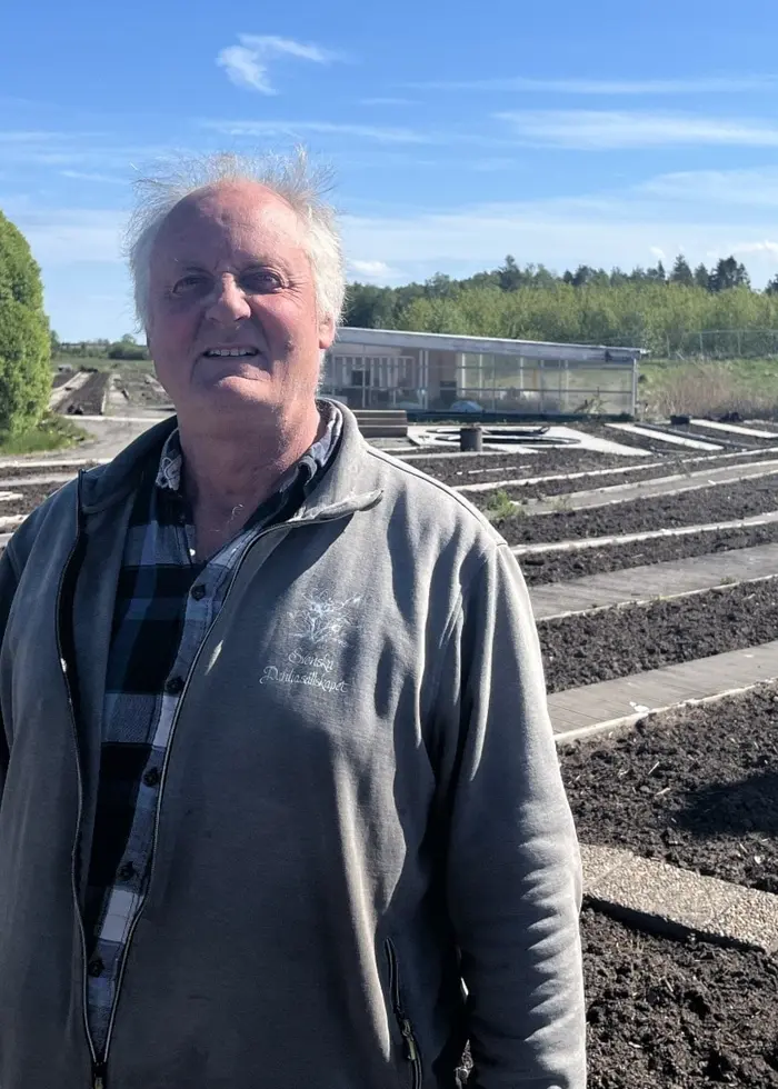 Blomsterodlaren Jan Ansbjer står utomhus vid en dahliaplantering utanför Nossebro, med långa odlingsrader och blå himmel i bakgrunden.