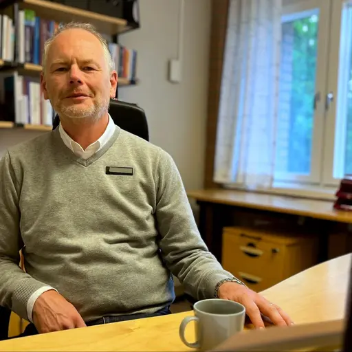 En man vid ett skrivbord på ett kontor. Han har en ljusgrön tröja över vit skjorta, tunt hår och framför honom står en kopp kaffe. På väggen bakom honom finns en bokhylla.