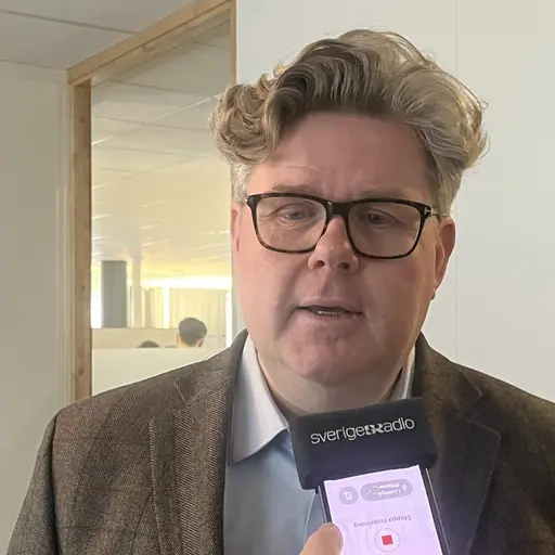 Justitieminister Gunnar Strömmer intervjuas inomhus, iförd kavaj och skjorta, med en mobiltelefon med mikrofon riktad mot sig i en kontorsmiljö.