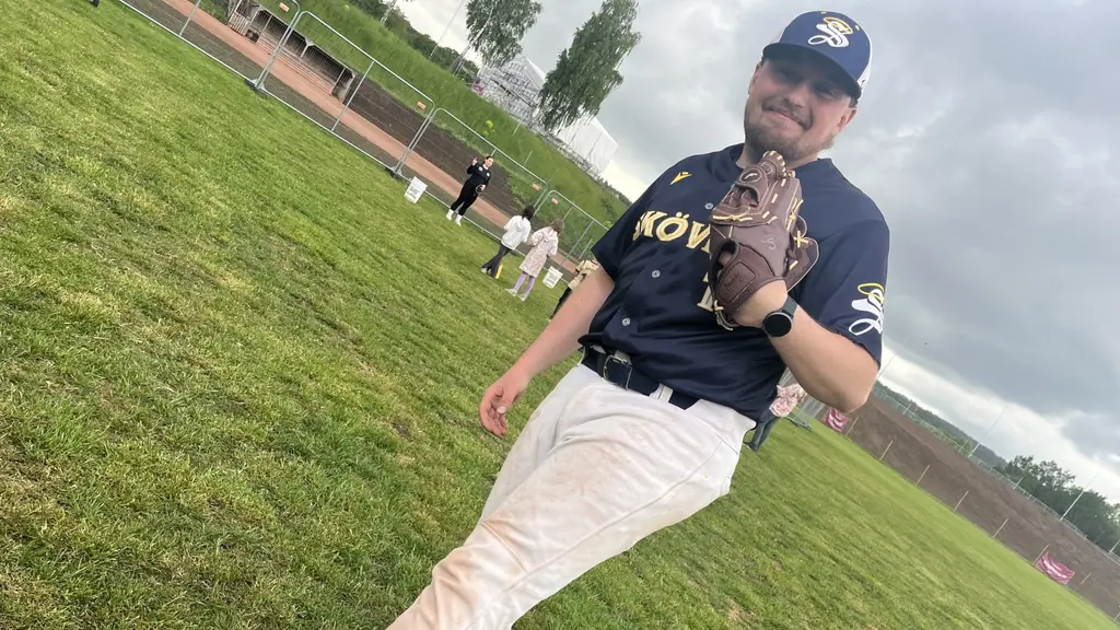 Baseballspelaren Adam Eklund i Skövde står på planen i mörk matchtröja och vit byxa, med handske framför ansiktet. Lagkamrater syns suddigt i bakgrunden.