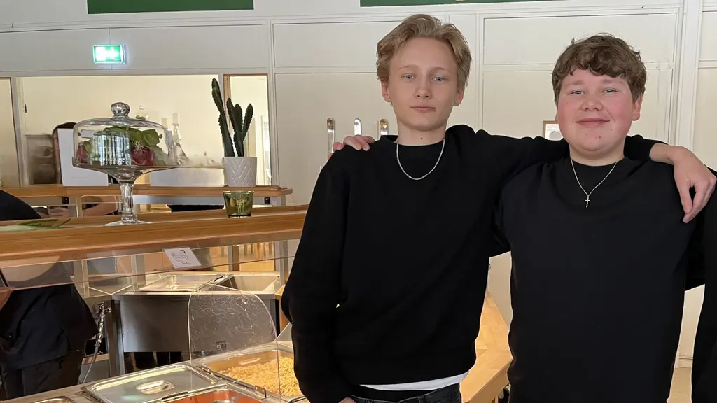 Cornelis Bornlid och Stig Jensen står vid en skolmatsal med en buffé fylld av olika maträtter, inklusive vegetariska alternativ. Bakgrunden visar en ljus och modern matsalsmiljö med dekorationer på väggarna.