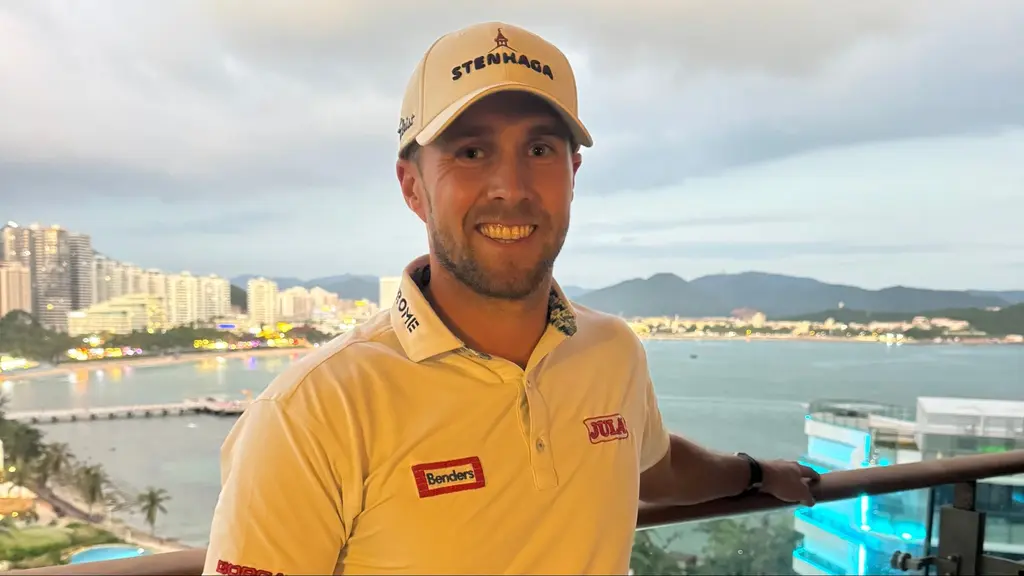 Golfaren Per Längfors från Tråvad står på en balkong med utsikt över en kuststad, höga byggnader och ett upplyst hav i bakgrunden. Han bär en vit keps och en vit golftröja med logotyper.