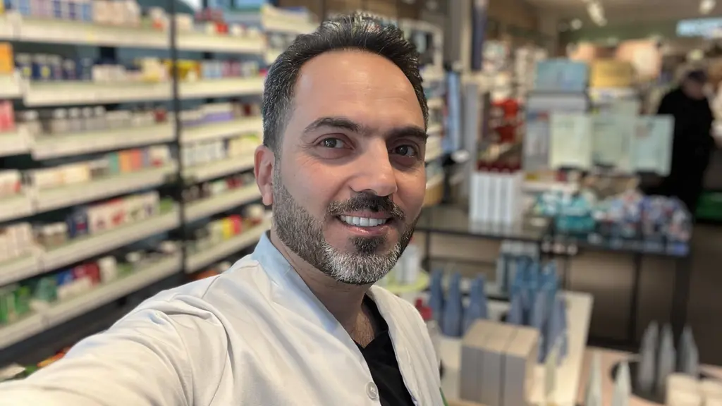 Mohamad Eiad Alhorani står i en butiksmiljö med apoteksprodukter på hyllor i bakgrunden. Han bär en vit arbetsrock som indikerar att han arbetar inom hälso- och sjukvård.