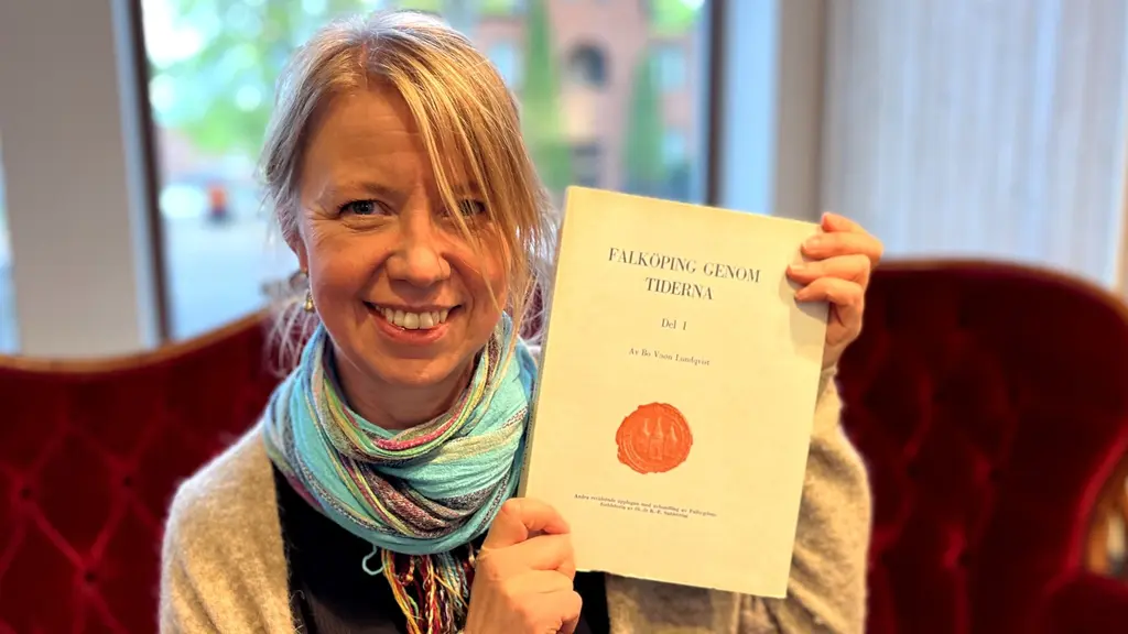 Ellinor Karlsson, chef på Falbygdens museum, håller upp en bok med stadsvapnet för Falbygden. Hon sitter i en röd soffa med ett fönster i bakgrunden som visar grönska utomhus. Ellinor har blont uppsatt hår, bär en beige kofta och en blåbrokig sjal.