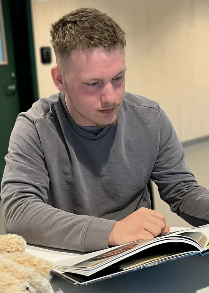 Pontus sitter vid ett bord och läser ur en barnbok. Bredvid den uppslagna boken ligger ett stort godsedjur.