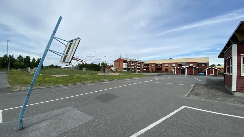 Prästholmsskolan i Boden med en blå basketställning på en asfalterad skolgård. I bakgrunden syns skolbyggnader med röda fasader och grönskande träd på avstånd under en molnig himmel.
