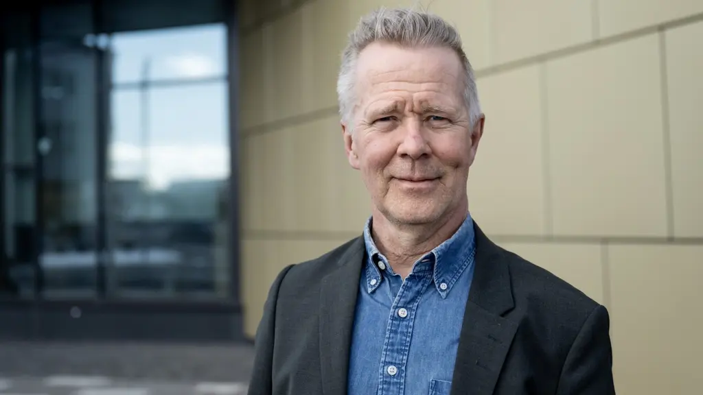 Mats Taaveniku (S), kommunstyrelsens ordförande i Kiruna, står inomhus i mörk kavaj och blå skjorta framför en modern vägg av ljusa paneler.