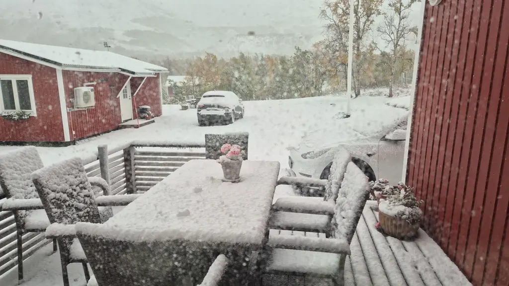Snötäckt trädgård i Björkliden med vita utemöbler, en bil och röda hus. Vinterlandskapets första snö har lagt sig på växter och möbler, vilket skapar en idyllisk och kall atmosfär.
