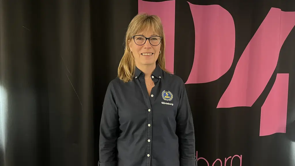 En person står framför en svart bakgrund med rosa och vita detaljer som visar logotypen för radiokanalen P4. Personen bär en mörk skjorta med ett tryck på bröstet.