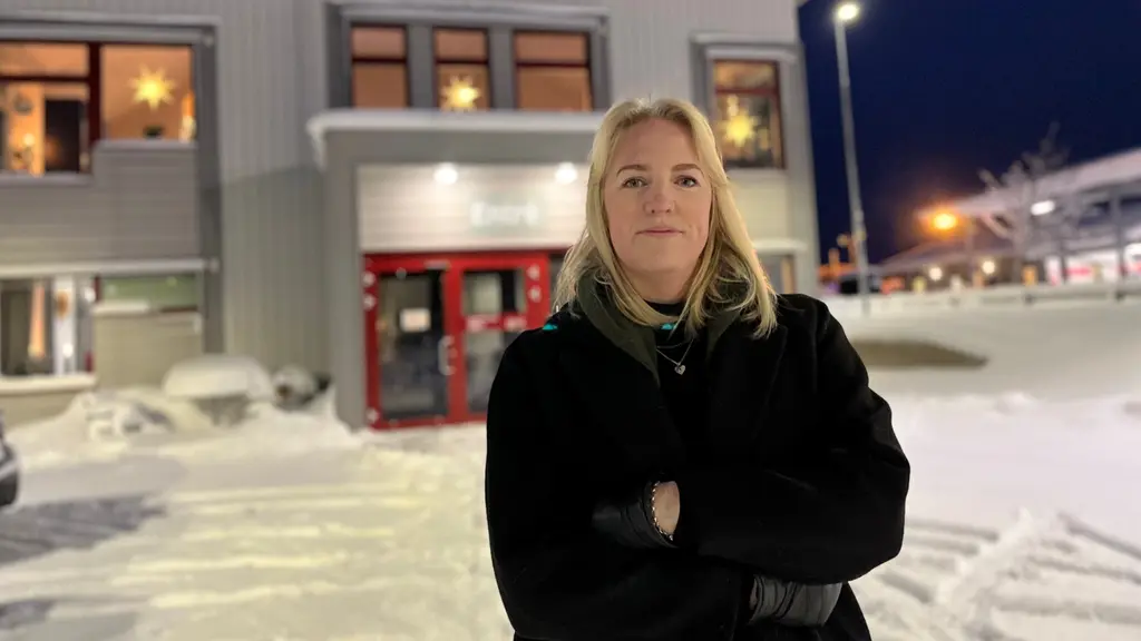 En person står utomhus framför en byggnad med stora fönster, dekorerade med stjärnor. Marken är täckt av snö och det är kvällsmörker. Byggnaden har röda dörrar och gatubelysning syns i bakgrunden.