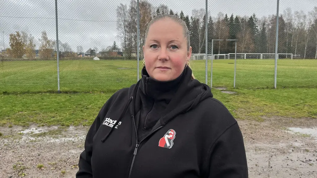 Ina Lööf står utomhus vid en gräsplan med fotbollsmål och höga nätstängsel i bakgrunden. Hon är klädd i en svart hoodie med innebandyrelaterad logotyp.