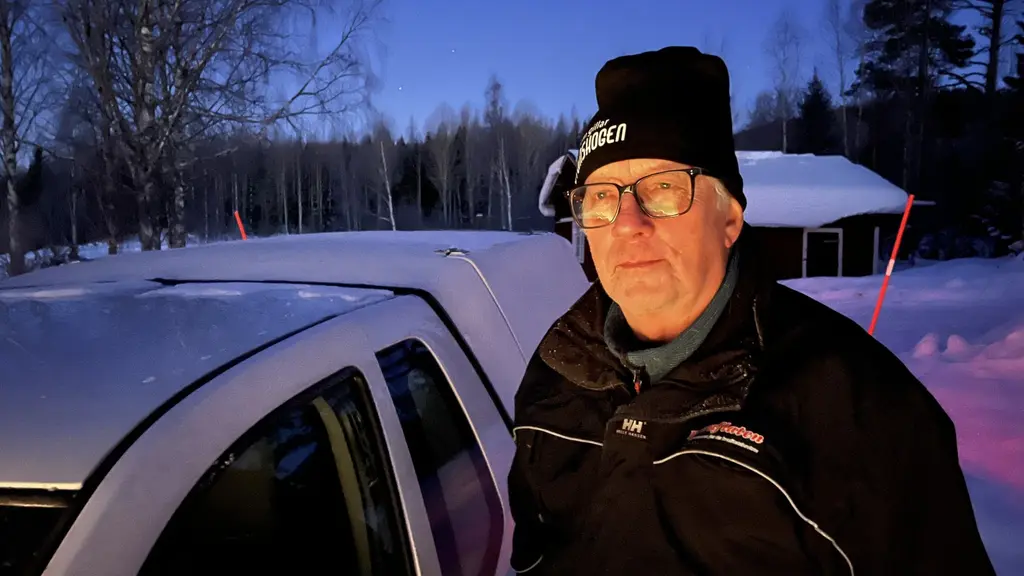 En person med vinterkläder står utomhus vid en snötäckt bil. I bakgrunden syns ett snölandskap med träd och en byggnad i skymningsljus.