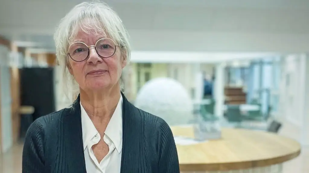En person står i en ljus kontorsmiljö med en rund receptionsdisk i bakgrunden. Personen är klädd i en vit skjorta och en mörk kofta.