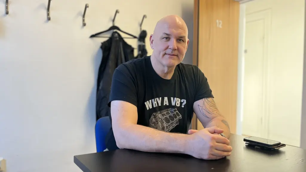En man sitter vid ett bord i ett rum med ljusa väggar och några jackor hängande på krokar i bakgrunden. Han har en svart t-shirt med ett tryck och en mobiltelefon ligger på bordet framför honom.
