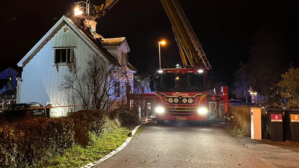 En brandbil står parkerad framför en villa i Bomhus under en nattinsats. Taket på villan är skadat och brandmän arbetar med en stegbil. Avspärrningar syns i området.