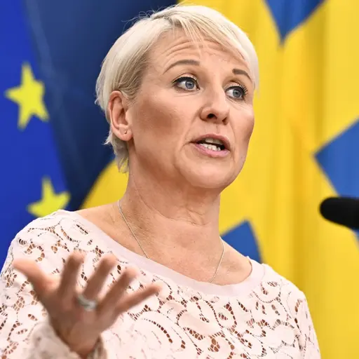 Äldreminister Anna Tenje (M) står vid en mikrofon under en pressträff med EU-flaggan och Sveriges flagga i bakgrunden. Hon bär en ljus topp och gestikulerar med ena handen.