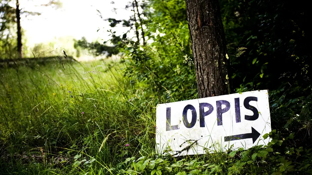 En handmålad skylt med texten 'Loppis' och en pil pekar mot höger, placerad vid en skogsdunge. Skylten signalerar en loppmarknad i lantlig miljö.
