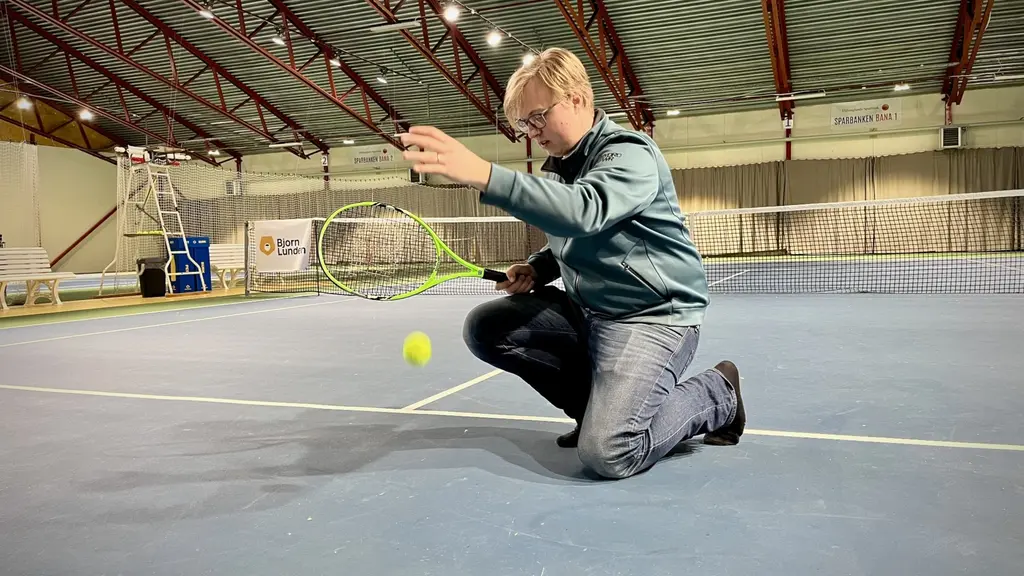 Björn Skyttberg demonstrerar studsproblem på en tennisbana inomhus. Han sitter på knä och håller en boll i handen, omgiven av nät och en välvd takstruktur.