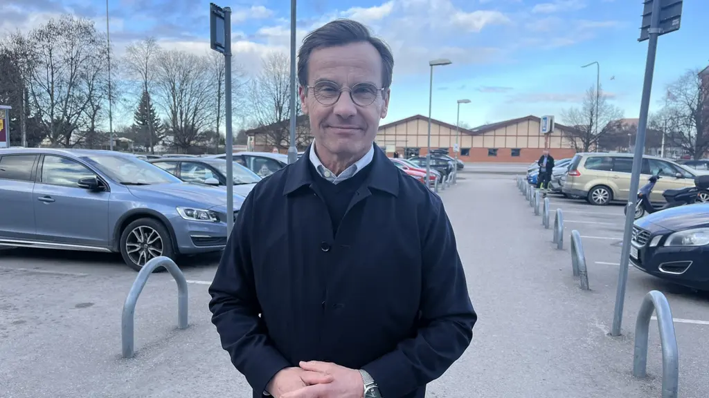 Statsminister Ulf Kristersson (M) står utomhus på en parkeringsplats med bilar i bakgrunden. Han är klädd i mörk jacka och håller händerna framför sig. Bakom honom syns en byggnad och blå himmel med moln.