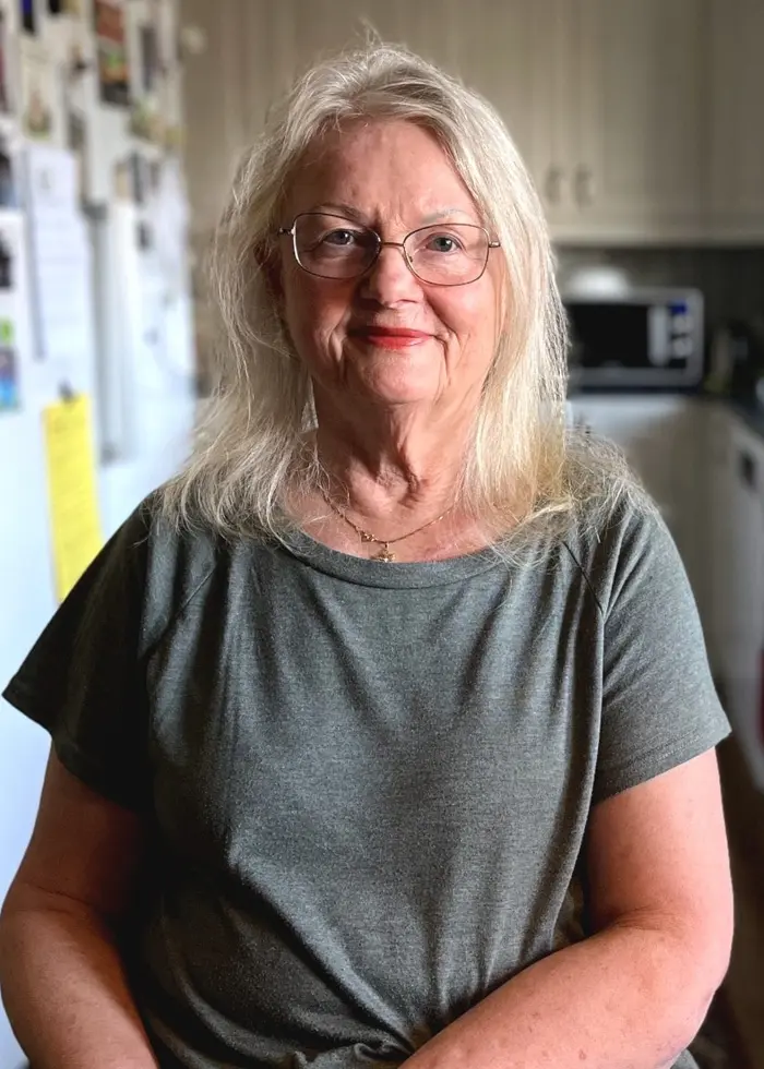 Kristina Grönvalls sitter i ett kök med ljusa skåp och moderna vitvaror. Hon bär en grön t-shirt och är fotograferad i hemmiljö. En köksstol syns i förgrunden.