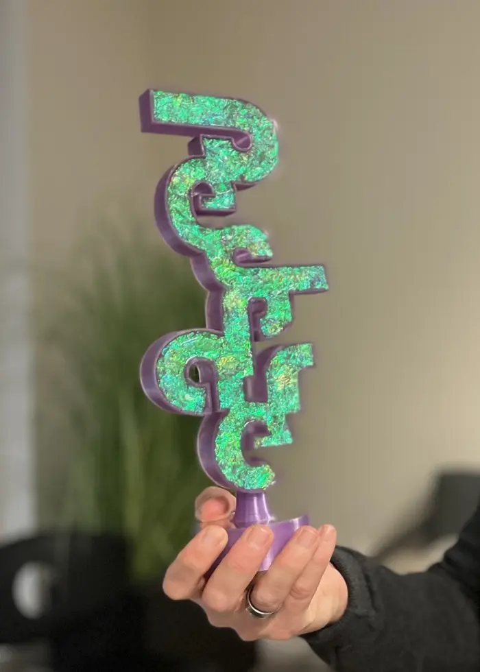 En person håller en färgglad statyett formad som text i en kontorsmiljö med suddiga väggar och en grön växt i bakgrunden.