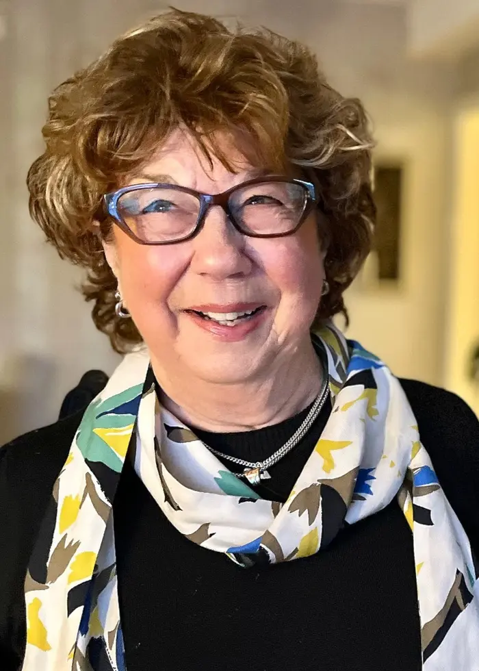 En person med lockigt hår står i ett ljust inrett rum med en vitrinskåp i bakgrunden. Personen bär en mönstrad halsduk och mörka kläder.