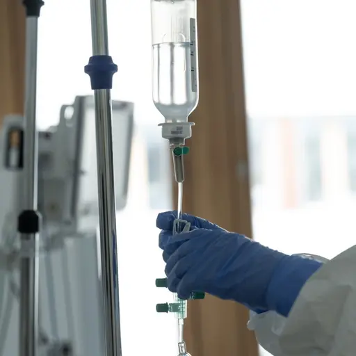 En sjuksköterska med blå handskar justerar ett dropp på en sjuksäng i en ljus sjukhusmiljö. Bilden är tagen på en canceravdelning vid Karolinska universitetssjukhuset i Solna.