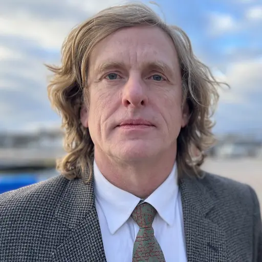 En person med långt blont hår bär en mönstrad kostym, vit skjorta och slips. Bakgrunden visar en utomhusmiljö med parkerade bilar och byggnader under en molnig himmel.