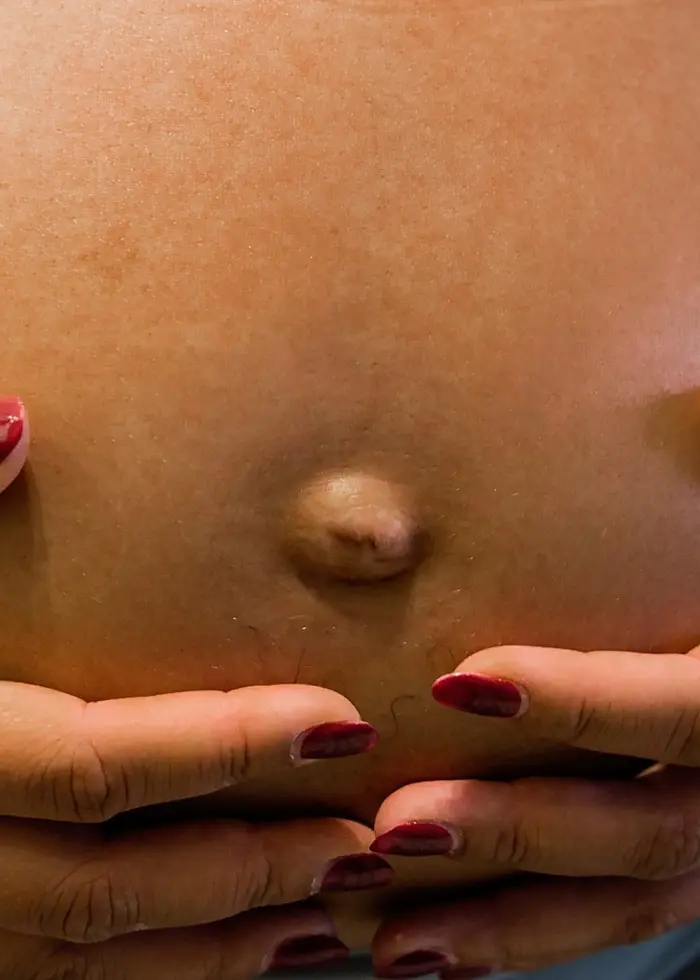 En gravid kvinna håller händerna om sin mage. Hon har rödmålade naglar. Fokus ligger på den runda magen, med en tydlig navel.