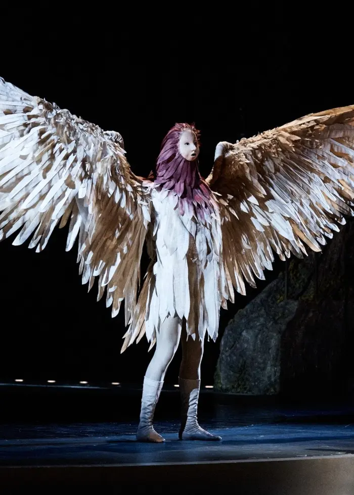 En scen från en operaproduktion där en person med stora, vita änglavingar står upplyst på scenen, medan en annan person sitter på golvet i en dramatisk position. Bakgrunden är mörk och mystisk.