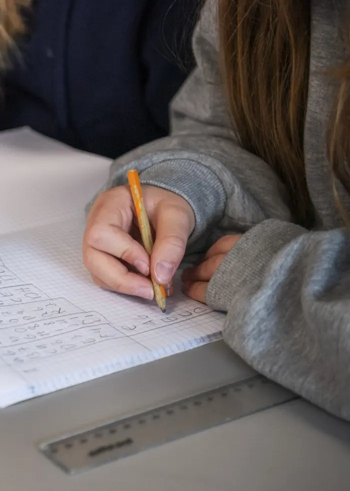 Elever sitter vid ett skrivbord i ett klassrum och arbetar med matematikuppgifter i sina skrivhäften. På bordet ligger en linjal, en matematikbok och olika skolmaterial.