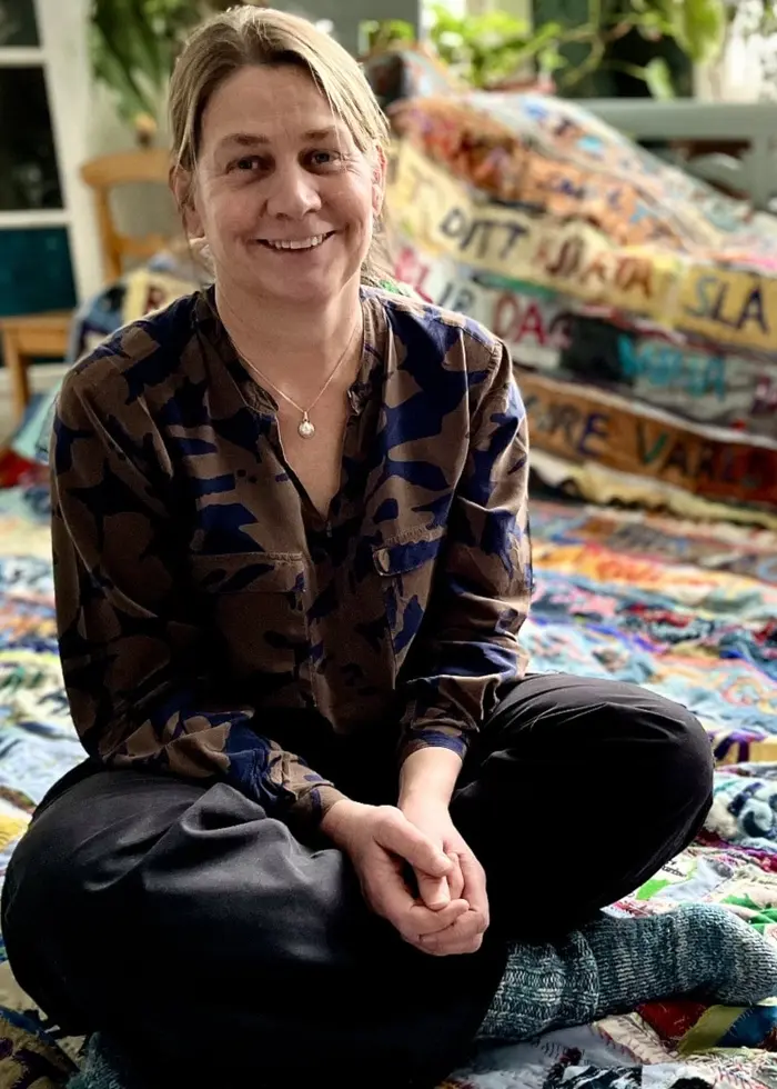 En person sitter på golvet i ett rum fyllt med färgglada textilier som har olika texter broderade på sig. Rummet är ljust med grönskande växter i bakgrunden.