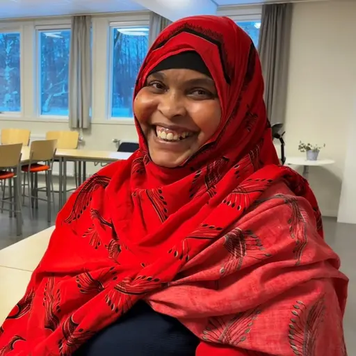 Maawerda Adam Omar klädd i en röd hijab sitter i ett klassrum med vita väggar, tomma stolar och bord. Ett stort fönster i bakgrunden släpper in dagsljus.
